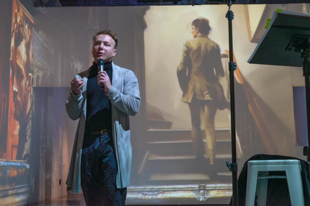 A man holds a microphone and speaks to an audience, dressed in a smart casual outfit with a coat and a scarf. He is standing in front of a projected image depicting a person ascending a staircase, which lends a dramatic and artistic tone to the scene. The speaker seems engaged and expressive, possibly in the midst of a lecture, presentation, or performance. The venue has the look of an art gallery or event space, and the large projection enhances the narrative or thematic elements of the speaker's presentation. A music stand with notes or a script is to his right, suggesting preparation and formality to the event.