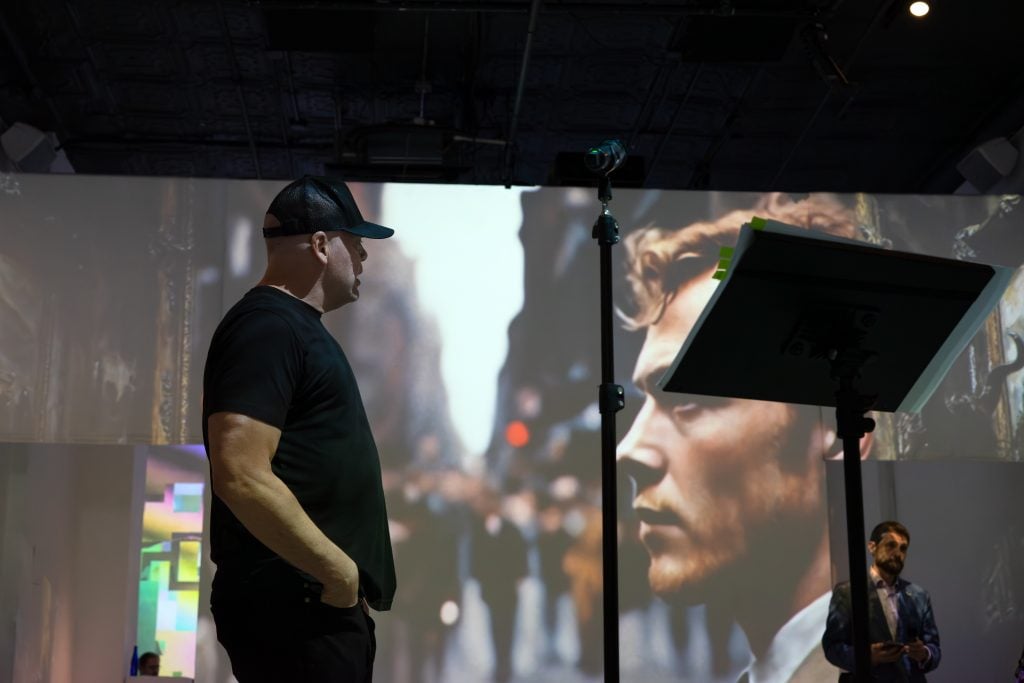In the foreground, a man wearing a dark t-shirt and a cap stands profile to the camera, appearing to address an audience or engage in performance. In the background, a large, detailed image is projected on a wall, featuring a close-up of a man's face, which contributes to the thoughtful atmosphere of the setting. Off to the right, another individual is partially visible, looking down at his smartphone, possibly awaiting his turn to speak or simply an attendee. The scene suggests an event focused on discussion, art, or performance, where visuals play a significant role in the experience.