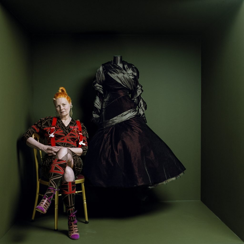 A woman in bright orange hair seated in a chair next to a dark satin ballgown on a mannequin