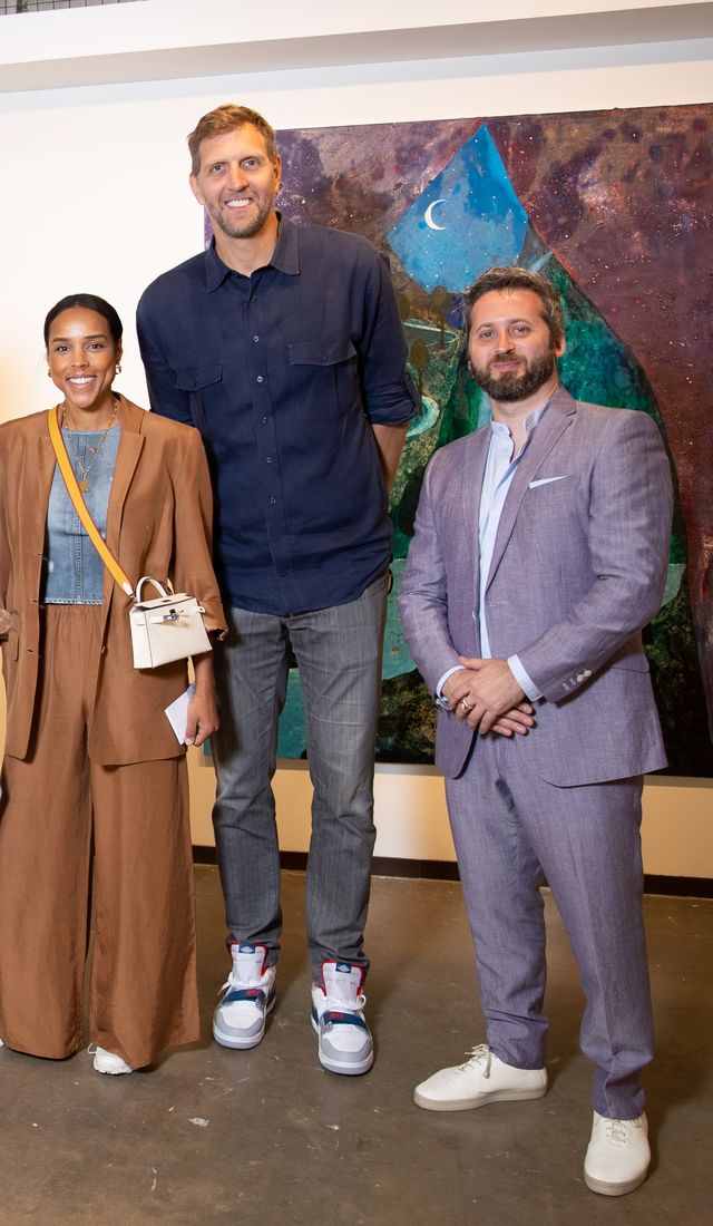 Three people pose for a color photo with a painting behind them—a woman, a very tall man, and a shorter man.