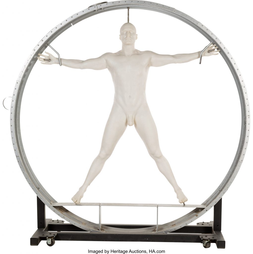 A photo of a sci fi prop featuring a white 3D printed male nude body suspended and spread within a grey metal ring