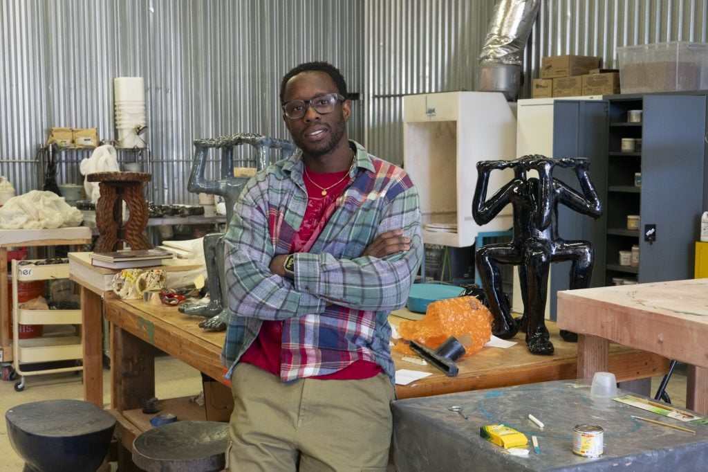 a young black man wearing glasses stands in an art studio with his arms crossed. sculptures are visible behind him