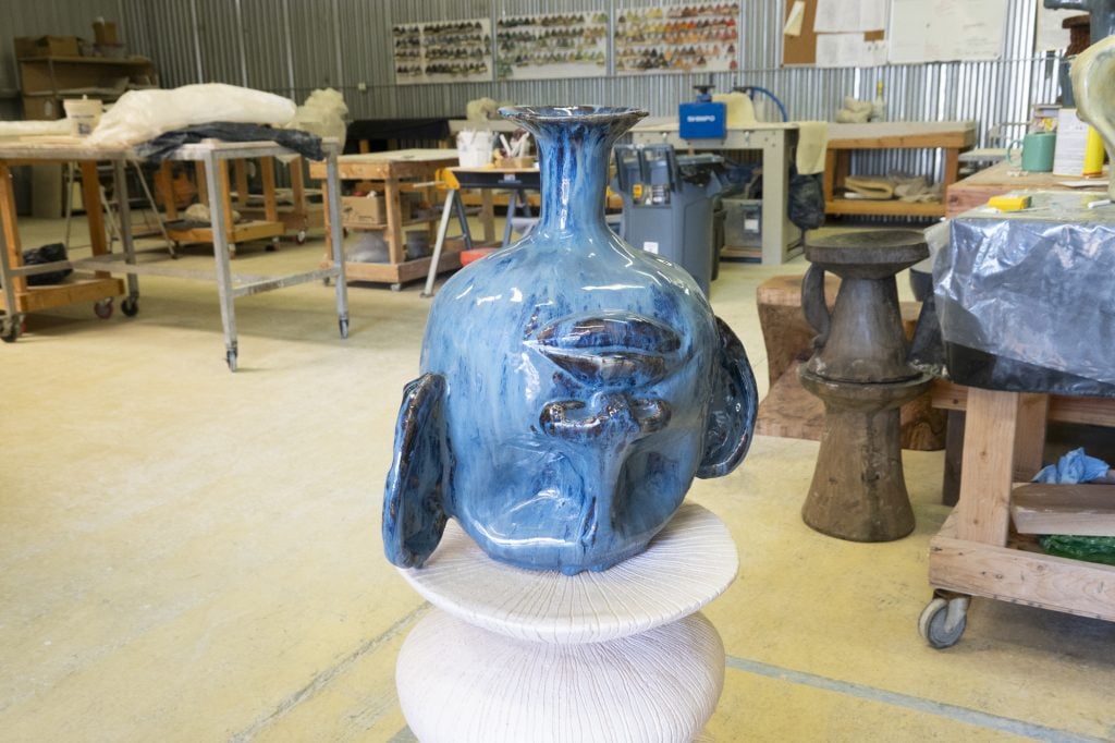 a ceramic sculpture, a vessel, showing a face with large ears, the face is upside down, and the entire vessel is a blue color