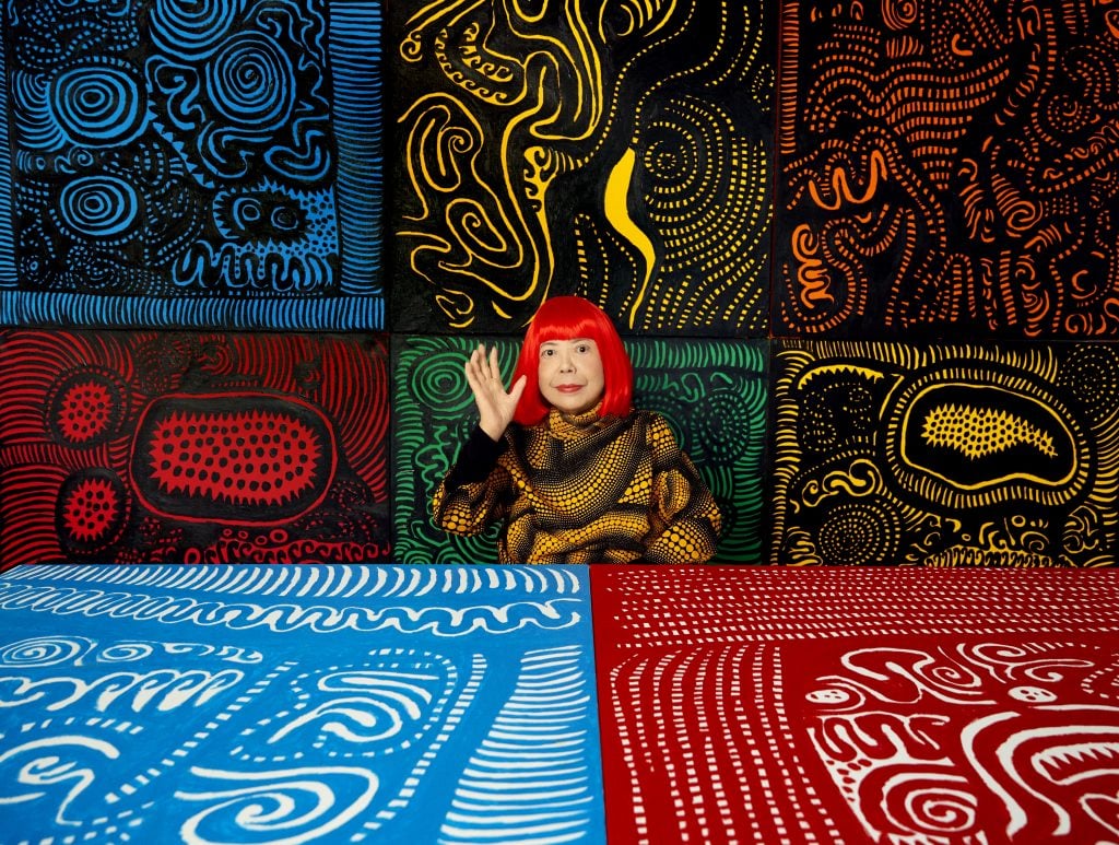 A woman, Japanese artist Yayoi Kusama, sitting a desk and in a room hung with her paintings of symbols