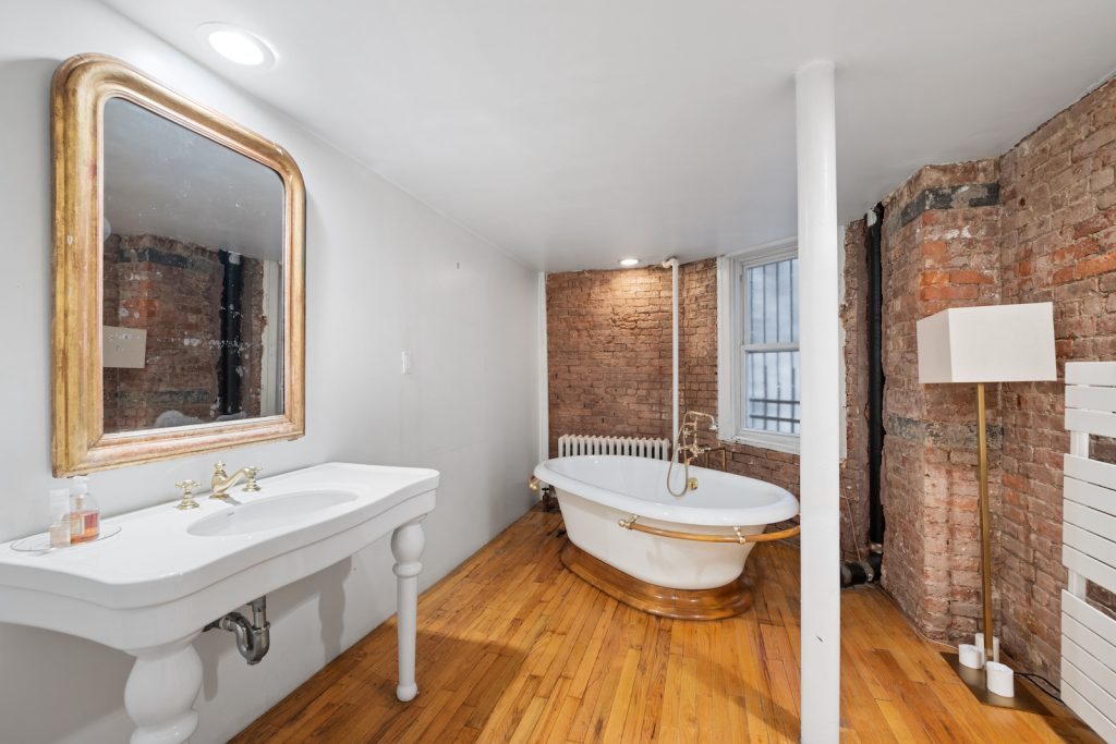 Inside a New York property, a white bathroom with a light brown hardwood floor. A roll top bath with gold features is in the centre, with a sink unit and gold mirror to the right. 