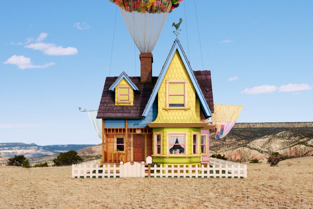 A multi-colored house, attached to a clump of balloons, sitting in a vacant lot