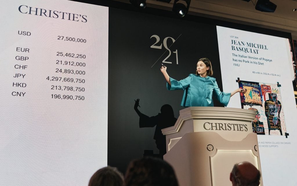 a woman in a blue suit stands at an auction podium and looks to the side of the room. the auction house is christies