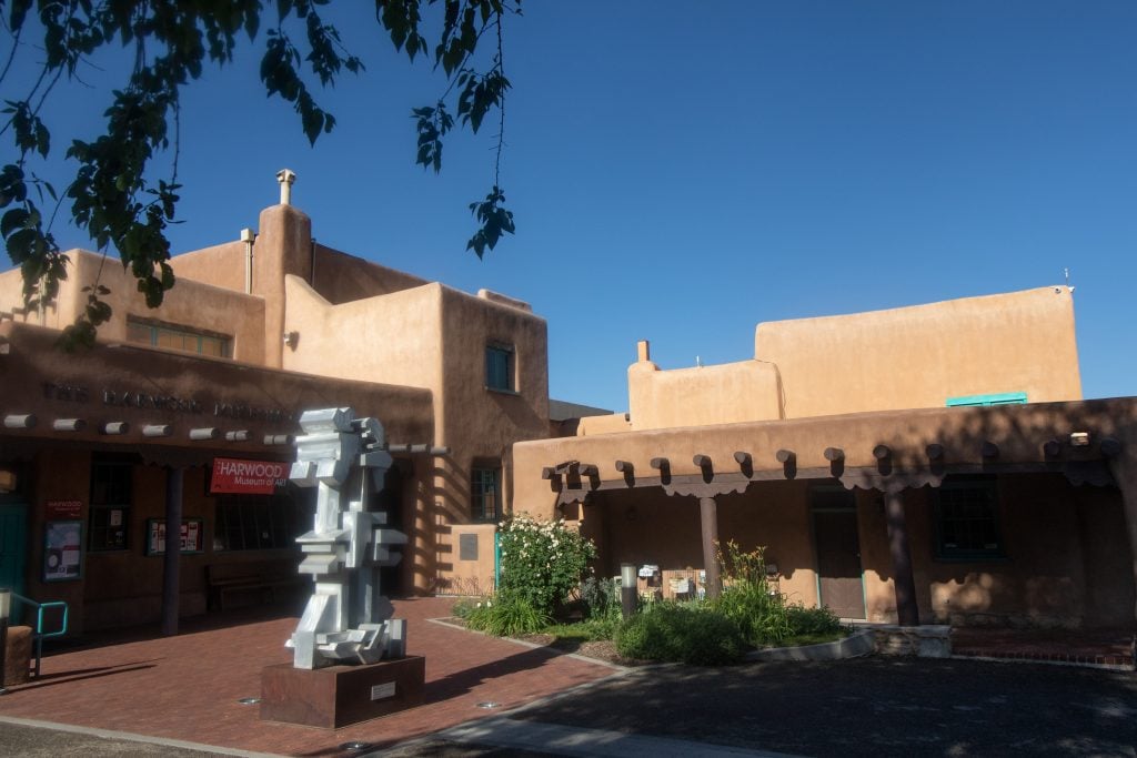  A daytime view of the Harwood Museum of Art, featuring its distinctive Pueblo-style architecture with smooth, earth-toned walls and rounded edges. In front of the museum, a modern abstract sculpture is prominently displayed on a pedestal. Lush greenery and clear blue skies complement the scene, creating a serene cultural atmosphere.