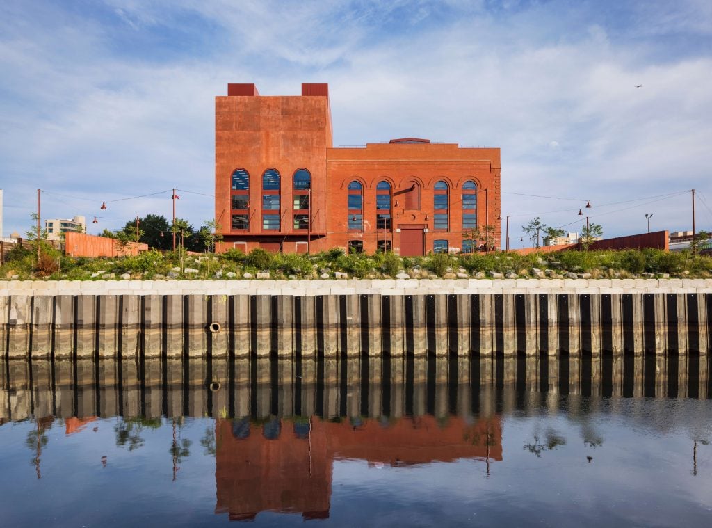 A large brick building with an industrial design is reflected in the calm water in front of it. The building features tall, arched windows and a rust-colored exterior. It is surrounded by a landscaped area with greenery and a sturdy waterfront retaining wall. The sky is clear with a few clouds, and the overall scene is tranquil and picturesque.