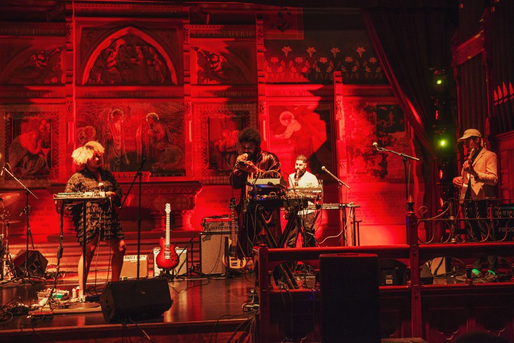 photograph of a four piece band perfroming on stage with biblical motifs in the background