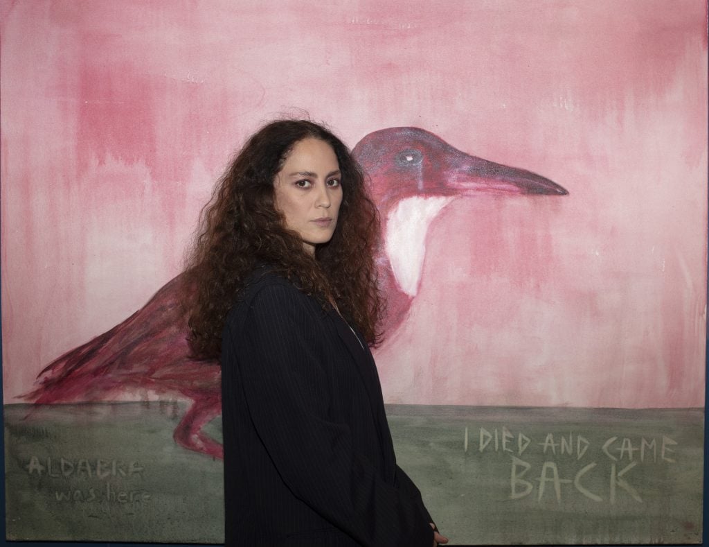 a woman with long curly brown hair stands in front of a painting with a big bird on it