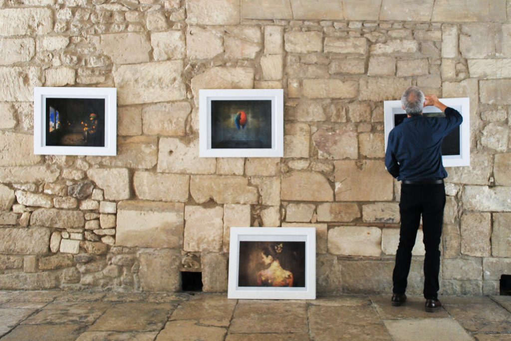 A man observes framed artworks displayed on an ancient stone wall inside a gallery. Three paintings are hung at eye level, and one is positioned on the floor against the wall. The visible artworks feature vibrant, somewhat abstract subjects, including a colorful bird and a figure, set against darker backgrounds, drawing attention to their details and colors.