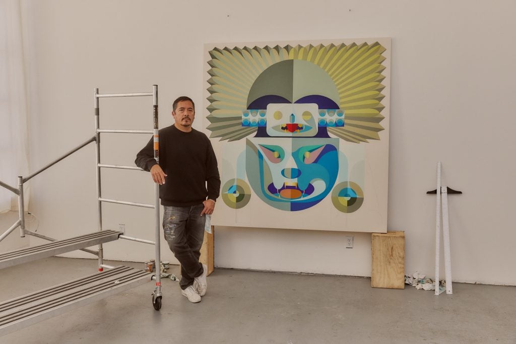 a man with short black hair wears a black sweatshirt and black jeans. He is standing with his arm resting on a studio scaffold, and in front of a brightly colored painting of a ritual mask
