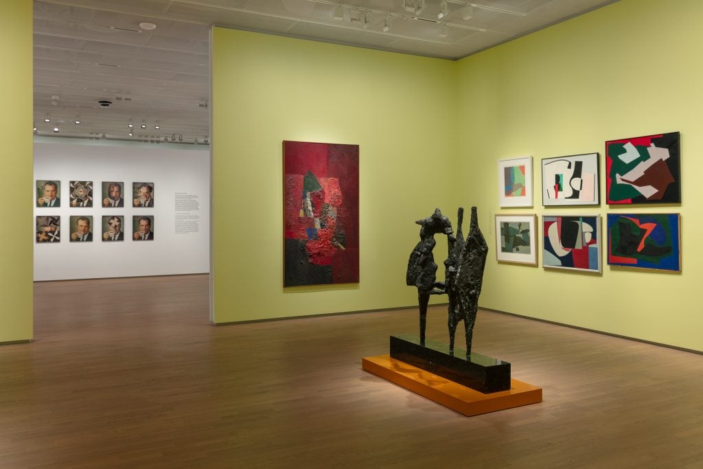 View of an art exhibition, with a dark colored bronze sculpture in the middle the room and many paintings on the walls.