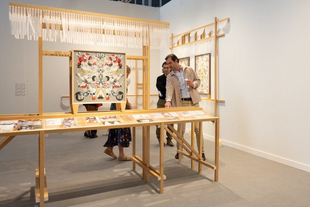 A color photograph shows a gray-walled art fair booth with intricate collage works and a table bearing different kinds of notes.