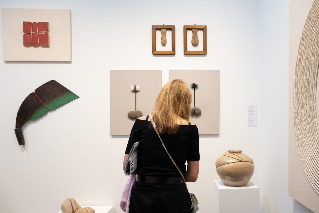 In a color photo, a person with a black dress and long hair stands in a white-walled booth displaying abstracts artworks, many of them bound with rope.