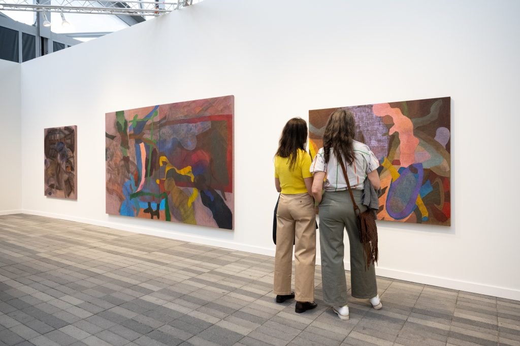 In a white-walled booth, two people examine colorful paintings in a color photo.