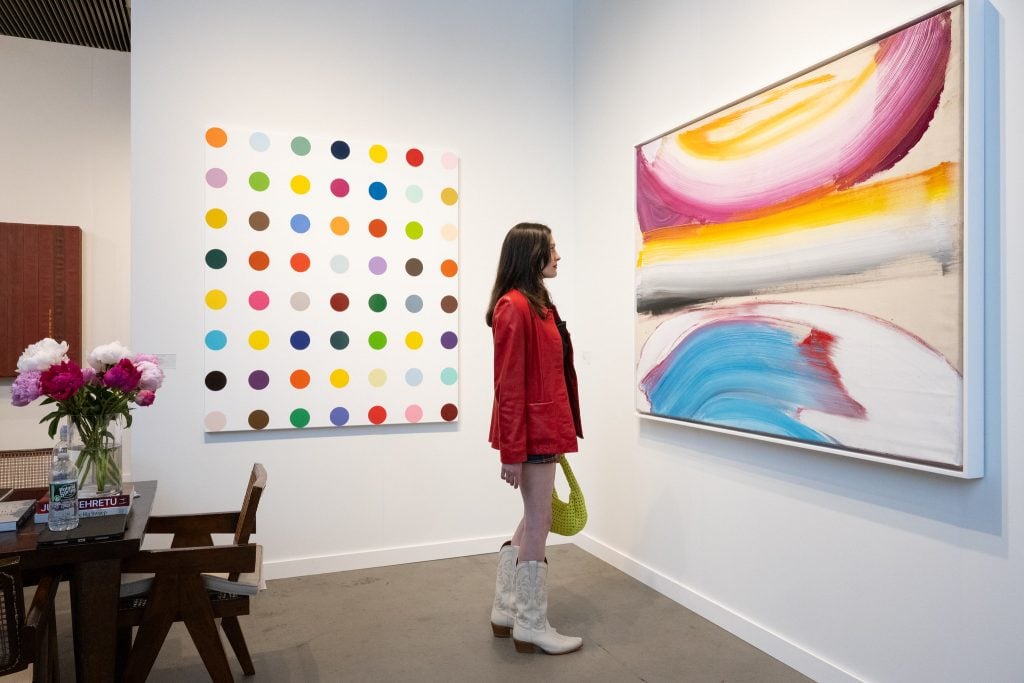 A lady wearing a red jacket and white cowboy boots stands in a white-walled booth with abstract paintings.