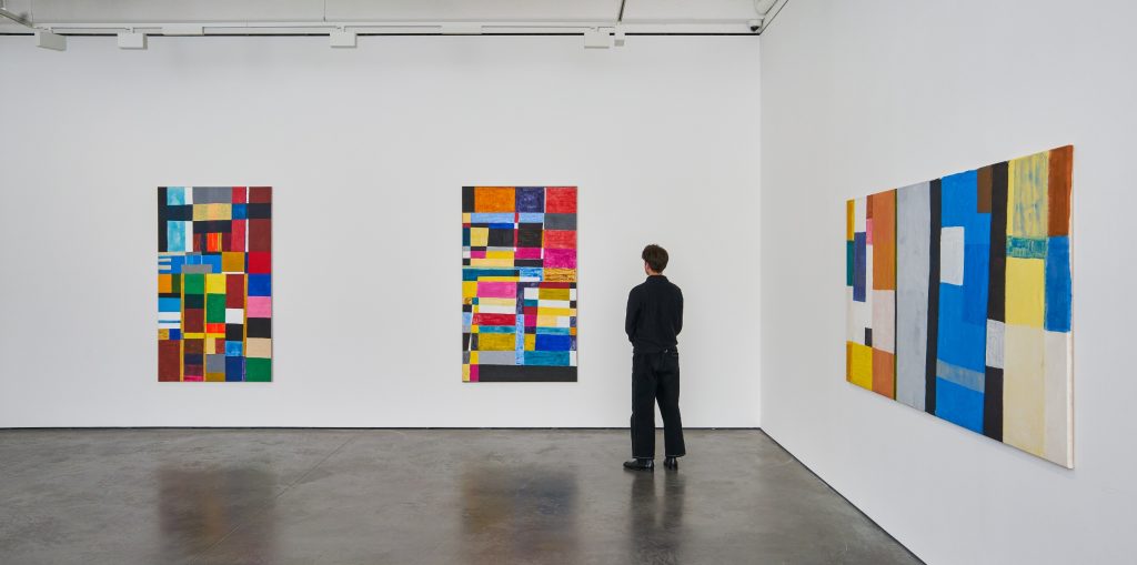 a white gallery space contains three paintings hanging on the wall that are composed of interlocking blocks of bright color, a man can be seen dressed in black looking at one