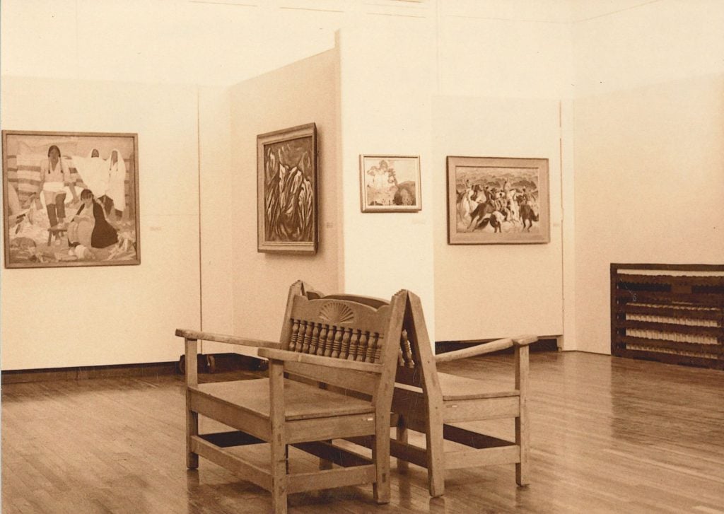 xt: An image of an art gallery interior with a wooden bench in the foreground. The walls display several paintings, including a large one with a group of people in a vivid, possibly outdoor scene, and other smaller framed artworks. The floor is wooden, and there's a decorative radiator cover on the right side. The overall atmosphere is quiet and contemplative.