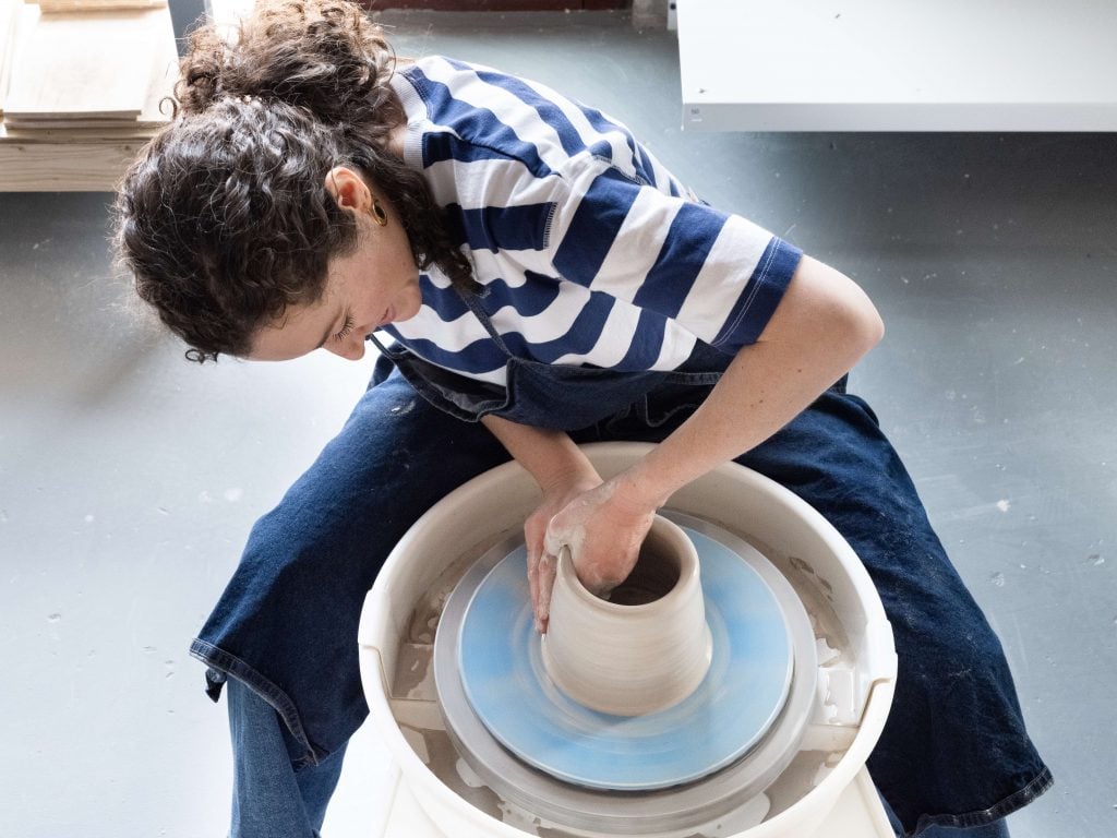 A person wearing a striped shirt and jeans is focused on shaping a clay pot on a pottery wheel. The workspace appears clean and well-organized, with additional materials stacked nearby. The individual is deeply engaged in the ceramic creation process, demonstrating skill and concentration.