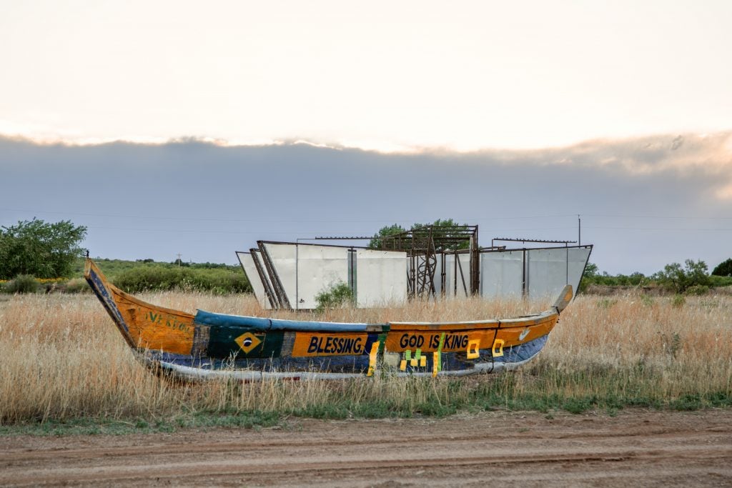 A painted boat with the words "BLESSING" and "GOD IS KING" sits in a grassy field, with a metal and glass structure in the background, under a cloudy sky.