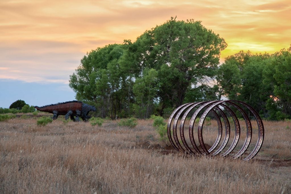 A field with metal circular sculptures in the foreground and a large, spiky dinosaur-like sculpture in the background, set against a backdrop of trees and a sunset sky.