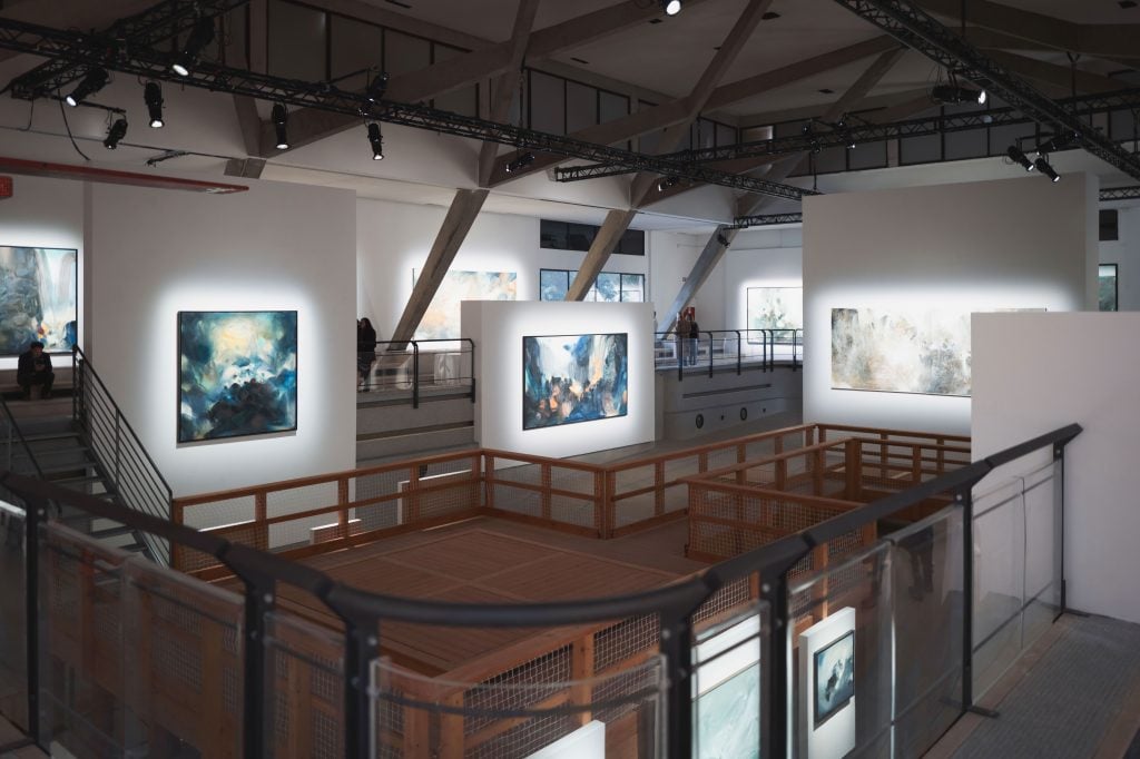 An exhibition of many paintings in a big space with high ceiling.