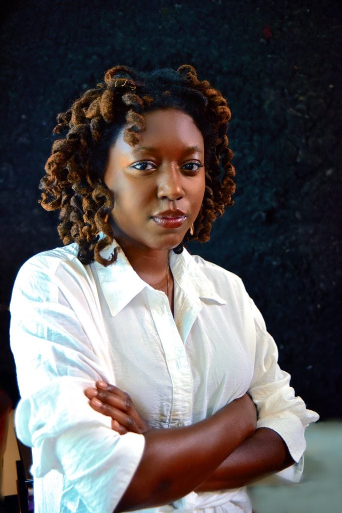 a photo of a young black woman in a white button-up shirt with her arms crossed. she looks directly at the camera