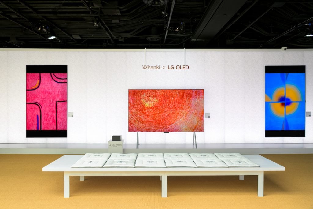 A room with three digital screens showing abstract paintings by Whanki. The central screen is red and yellow, the left pink and black and the right yellow and blue. 