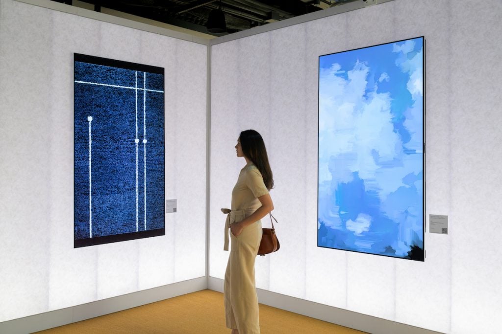 A brunette woman in a cream jumpsuit looks at two LED screen showing blue abstract art by Kim Whanki.