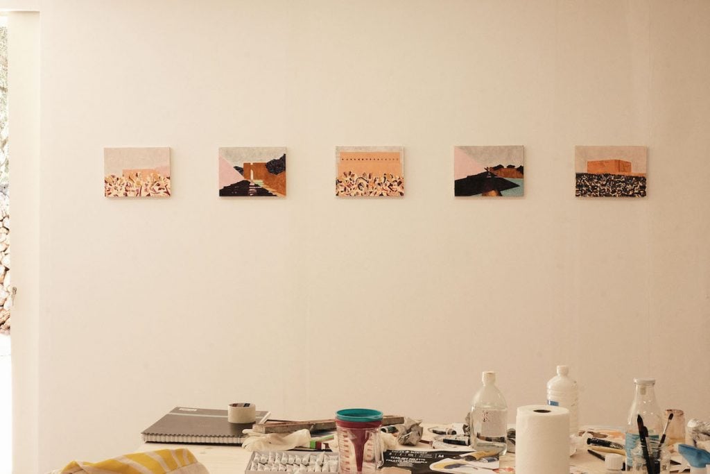 Five small paintings hang in a line on a pale wall, with a table of art supplies in front of them