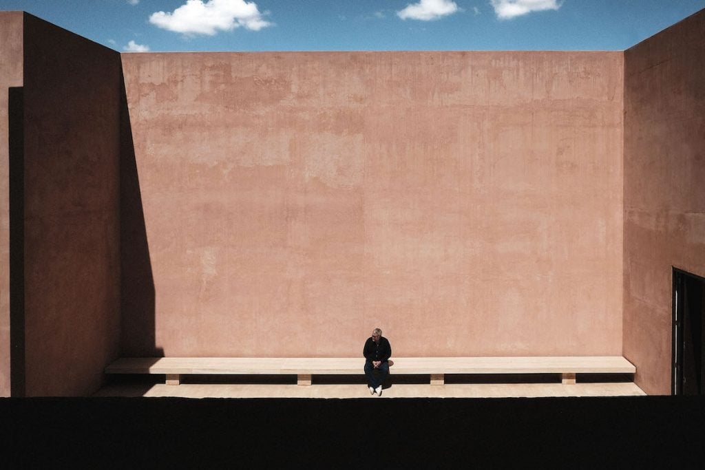 a man sits on a bench surrounded by tall terracotta walls, with the blue sky above