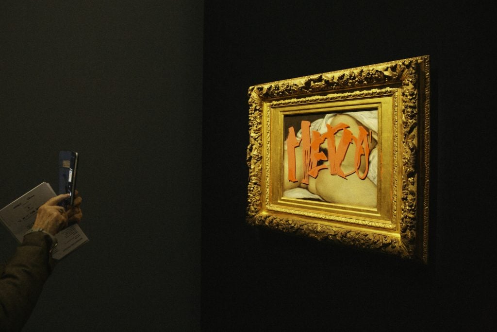 A painting of a nude woman in a gold frame tagged with the words 'Me Too' written in red paint. A viewer on the left takes a photo with their phone