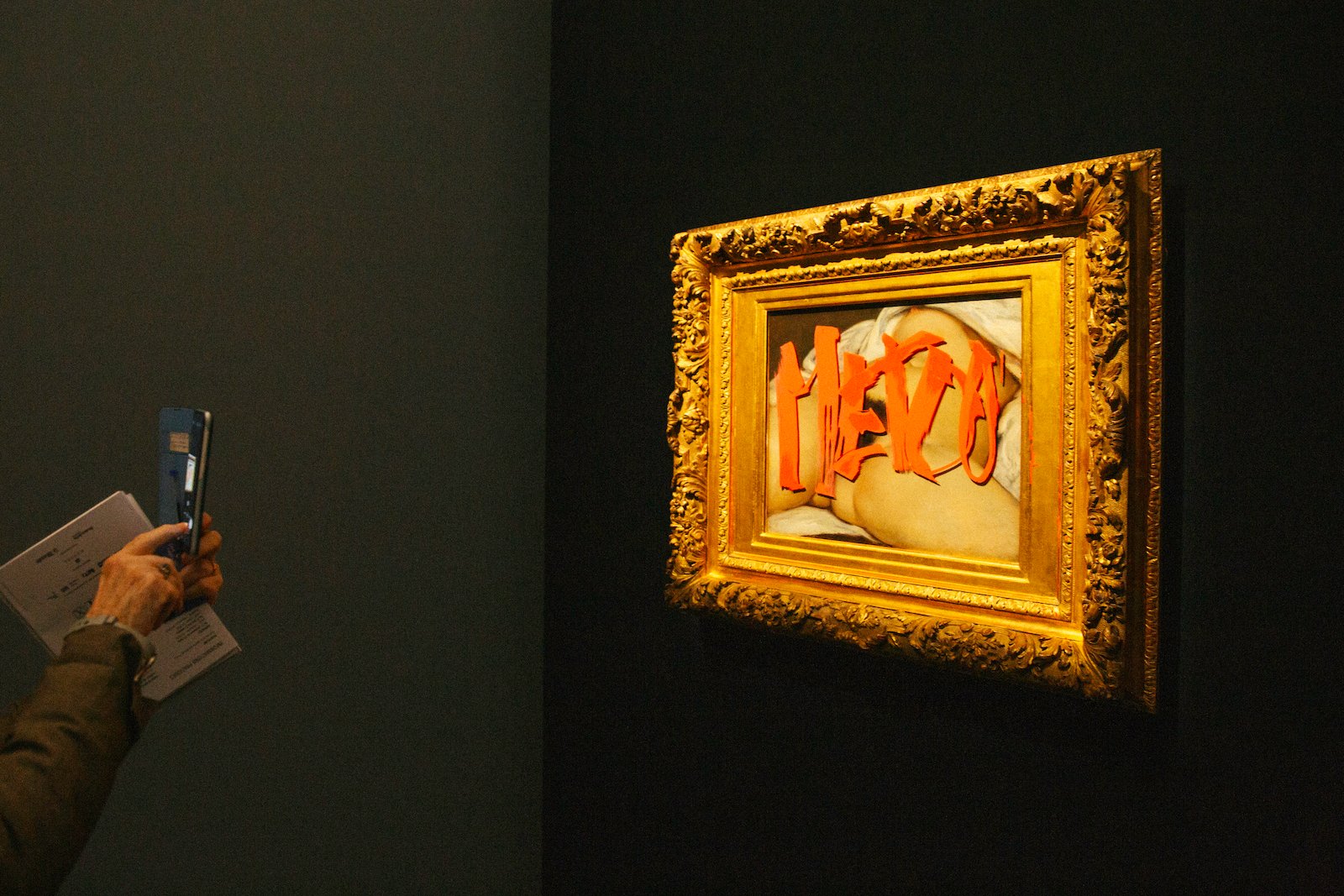 A painting of a nude woman in a gold frame tagged with the words 'Me Too' written in red paint. A viewer on the left takes a photo with their phone