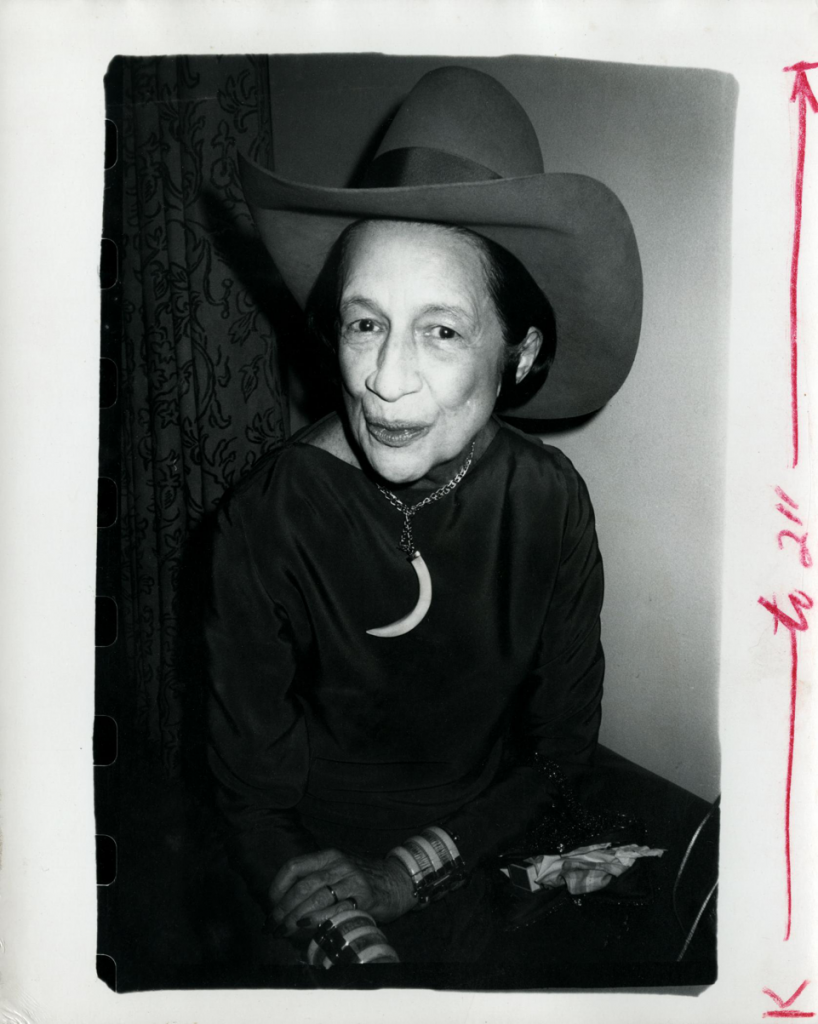 a silver print of a woman diana vreeland with a coy smile wearing a cowboy hat and a half moon bone necklace