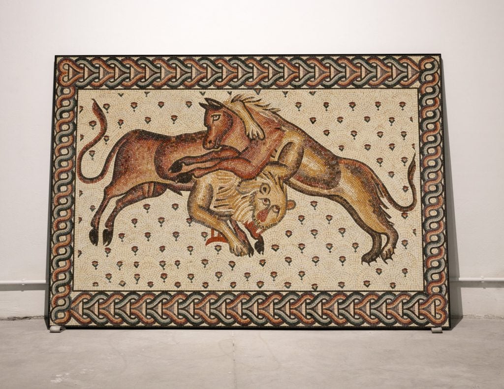 A mosaic depicting a dynamic scene of a lion attacking a bull, framed within a decorative border featuring intricate knotwork and geometric patterns. The artwork is rich in detail and uses a variety of earth tones.