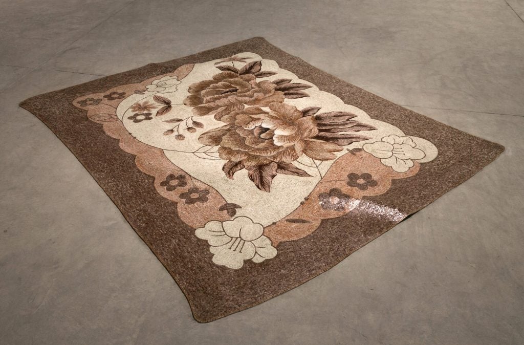 A mosaic rug on a gallery floor, intricately designed with floral patterns in natural brown and beige tones, featuring large blossoms and surrounding smaller floral motifs.