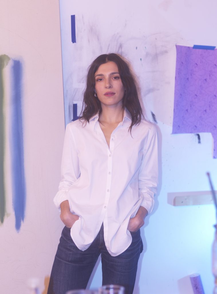 A brunette woman wearing a white shirt and jeans stands in front of a n unfinished painting in shades of white and blue
