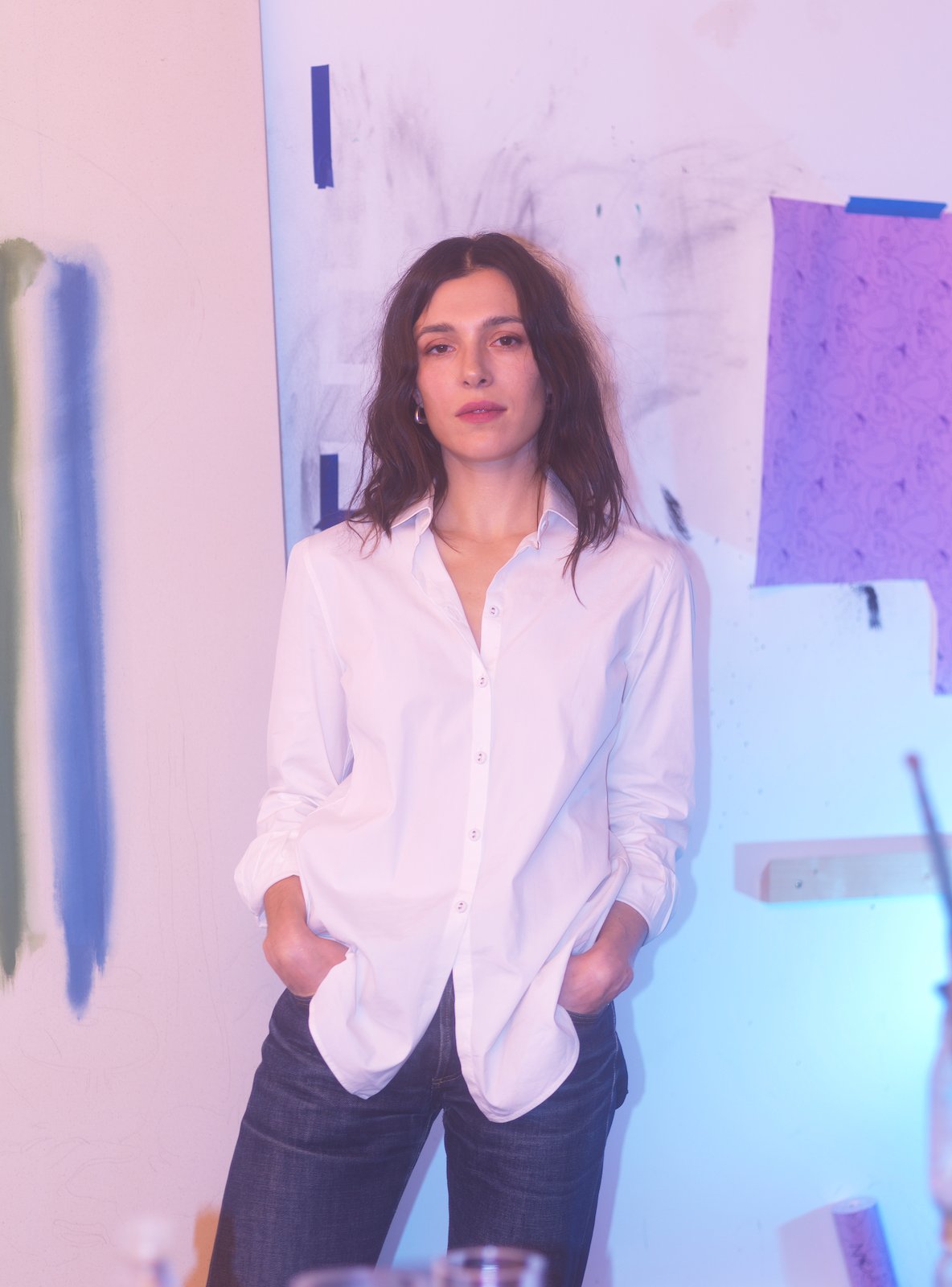 A brunette woman wearing a white shirt and jeans stands in front of a n unfinished painting in shades of white and blue