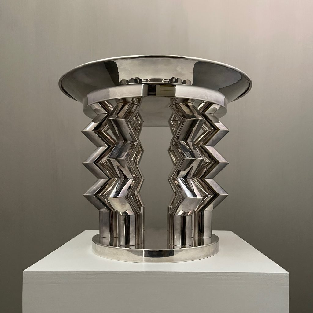 a metal chalice with zig zag legs sits atop a plinth by the designer Ettore Sottsass
