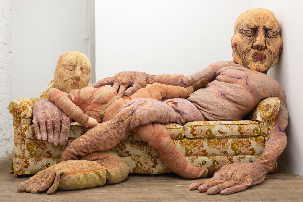 two enormous reclining knitted figures ae on an ugly 1970s couch 