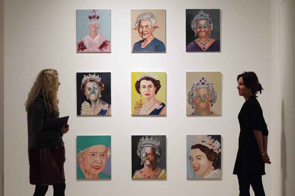 a grid of nine small portrait paintings hanging on a wall with two people standing to the side looking at them 