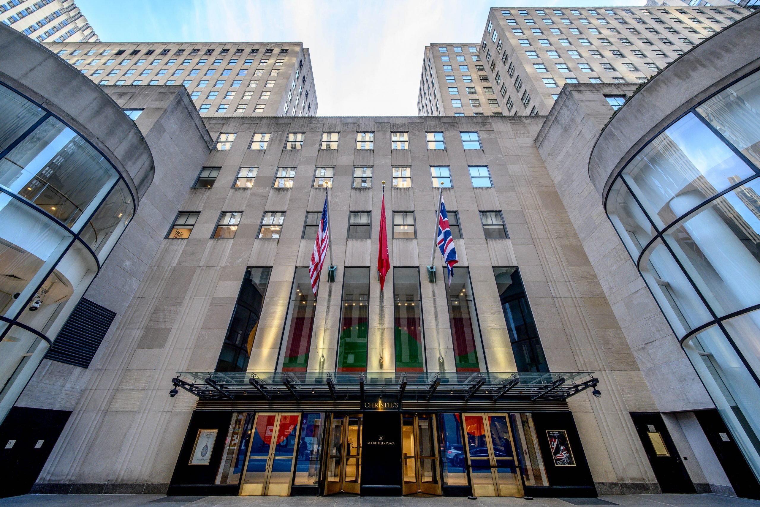 A color photo looks up at the facade of Christie's imposing headquarters in New York. There are many glass doors and flags flying above them.