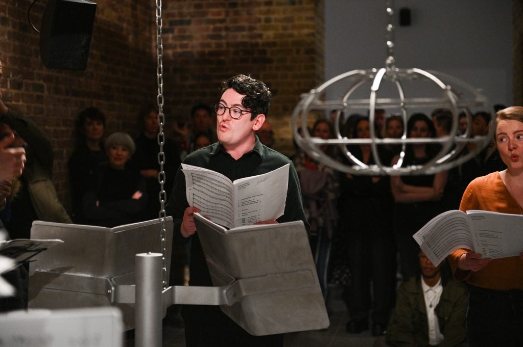 a man is seen reading from sheet music and behind him a crowd is watching, in the foreground are suspended open pieces of sheet music that are part of a larger installation that is hung from the ceiling