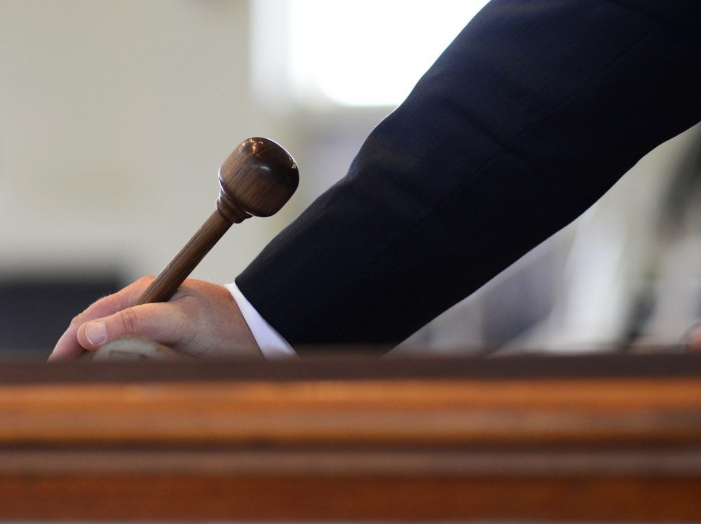 A color photo shows a besuited hand holding a wooden gavel atop a wooden table of some sort.