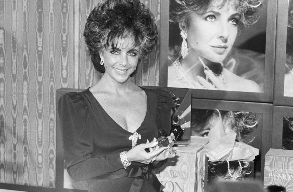 a smizing Elizabeth Taylor wearing a feather pin holds up a bottle of perfume