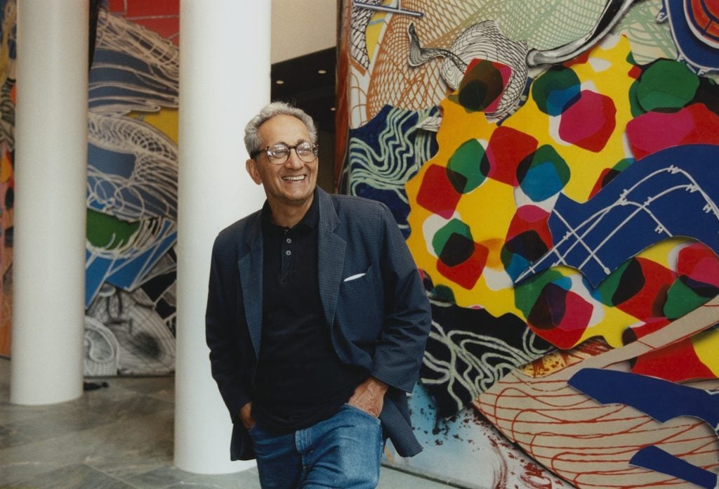 A middle-aged man, the artist Frank Stella, in a suit jacket stands in front of huge abstract paintings in a color photo