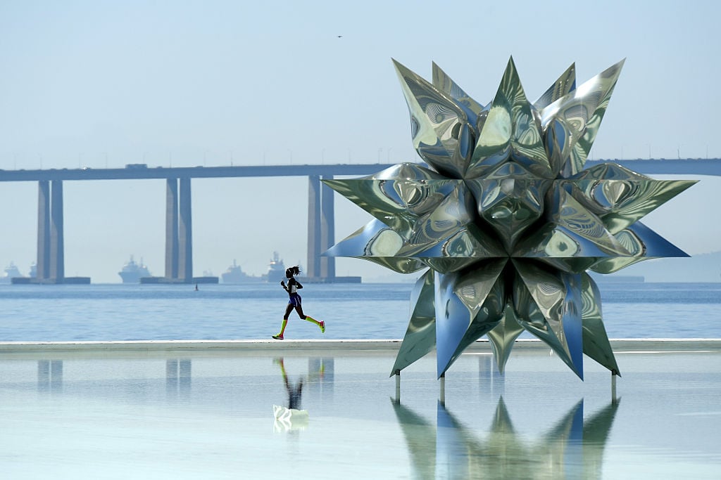 A woman in athletic gears runs in a color photo behind a huge sculpture of a steel star. The sea, and ships, and a highway are behind here.