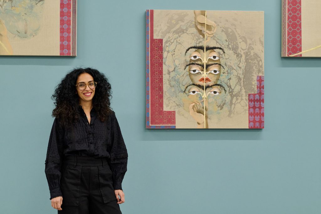 a women in a black dress with long black curly hair and glasses stands beside a painting of a surrealist face with many sets of eyes and eyebrows